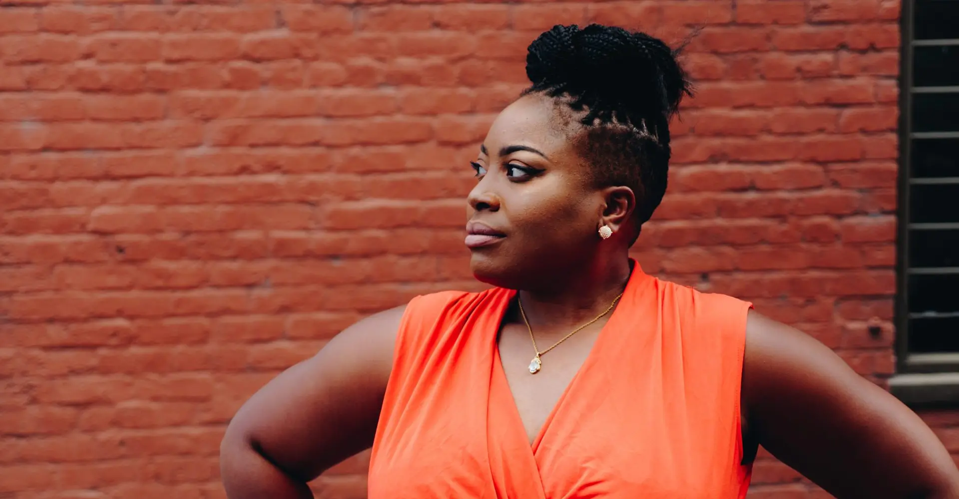 african-american-woman-in-orange-top-standing-proudly-with-hands-on-hips-against-brick-wall