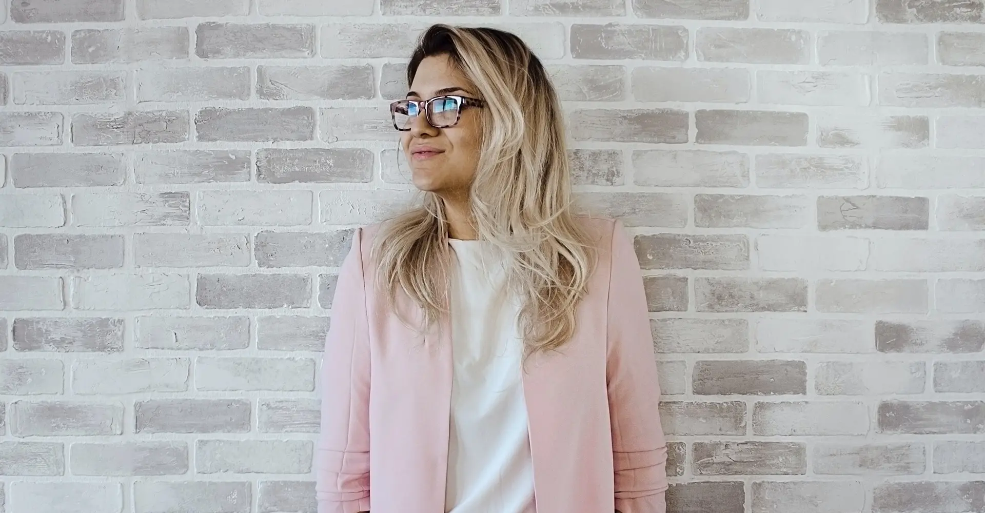 millennial-woman-with-blonde-hair-wearing-pink-jacket-leaning-against-light-brick-wall